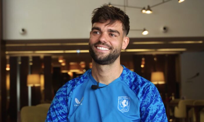 Robin Pröpper smiles during an interview while wearing an FC Twente training top, seated indoors with warm lighting in the background.