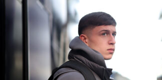 Young footballer Mikey Moore wearing a grey puffer jacket and backpack, looking focused as he stands outdoors beside a reflective surface.