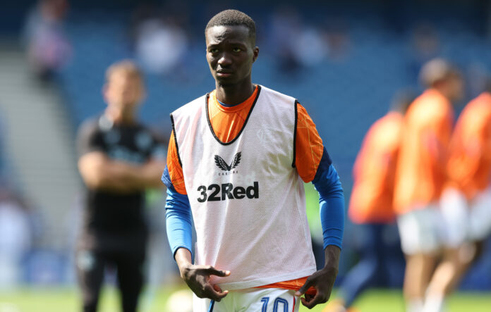 Mohamed Diomande warming up in a Rangers training vest during a pre-match session at Ibrox Stadium, with teammates and staff blurred in the background.