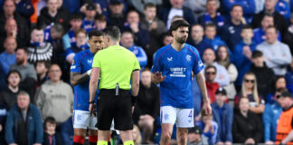 Rangers FC players James Tavernier and Robin Propper speak to the referee during a UEFA Europa League 2024/25 quarter-final match at Ibrox Stadium, with a focused crowd watching in the background.