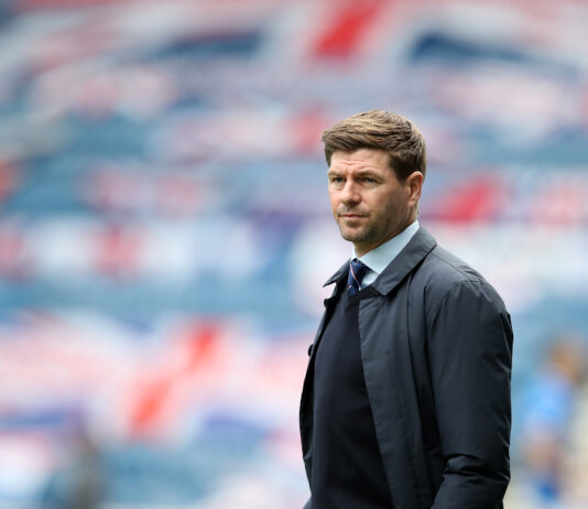 Steven Gerrard standing on the touchline in a dark coat and tie, with a blurred Union Jack display in the background at Ibrox Stadium.