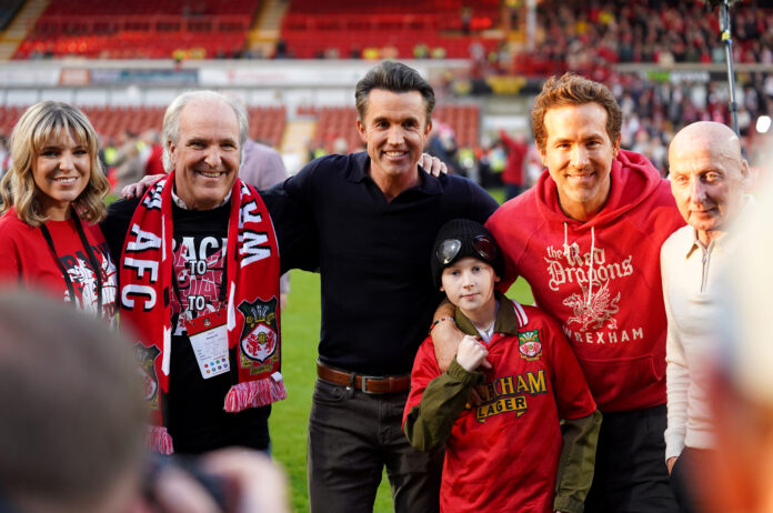Wrexham AFC co-owners and fans, including Rob McElhenney and Ryan Reynolds, pose for a celebratory photo on the pitch with supporters wearing club gear.