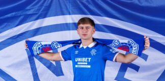Mikey Moore poses in a Rangers shirt holding a blue Rangers scarf above his shoulders in front of a large Rangers FC banner.