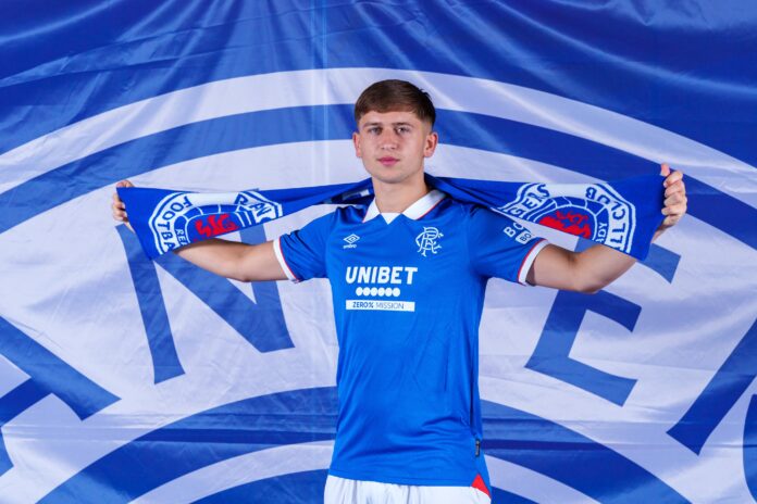 Mikey Moore Unveiled as Rangers’ Newest Signing Mikey Moore poses in a Rangers shirt holding a blue Rangers scarf above his shoulders in front of a large Rangers FC banner.