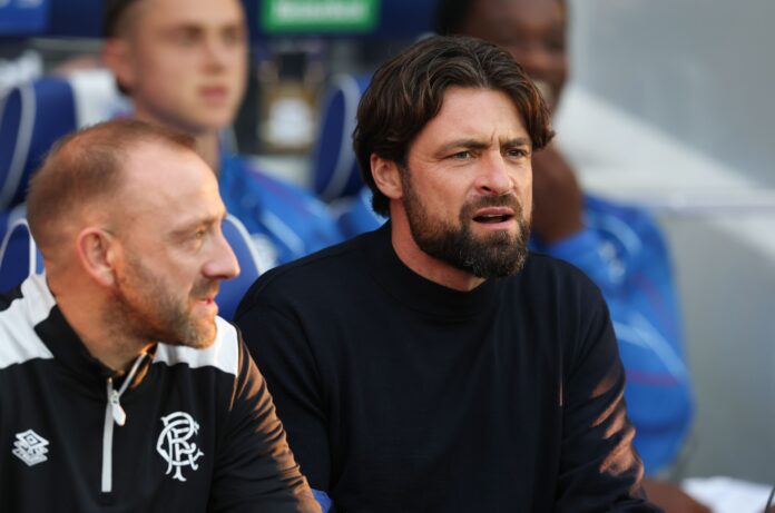 Rangers manager Russell Martin on the bench in a black jumper, looking concerned during the UEFA Champions League play-off first leg against Club Brugge, seated beside a staff member in Rangers training gear.