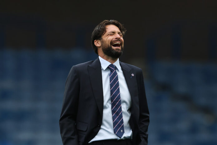 Russell Martin, dressed in a navy suit and striped Rangers tie, appears frustrated or shouting during a UEFA Champions League match, with empty blue stadium seats blurred in the background.