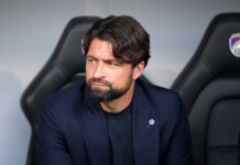 Russell Martin sitting in the dugout during a match, wearing a navy blazer and black shirt, looking to the side with a serious expression.