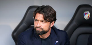 Russell Martin sitting in the dugout during a match, wearing a navy blazer and black shirt, looking to the side with a serious expression.