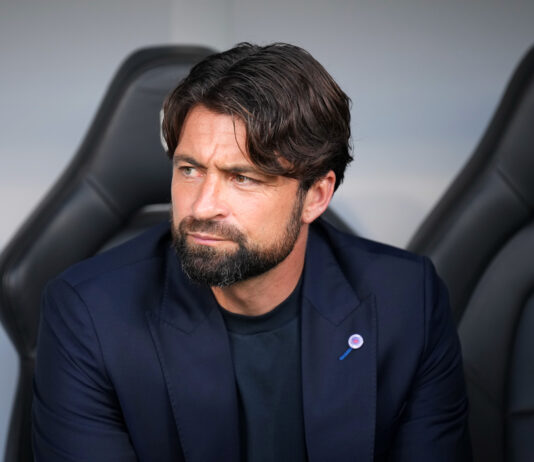 Russell Martin sitting in the dugout during a match, wearing a navy blazer and black shirt, looking to the side with a serious expression.