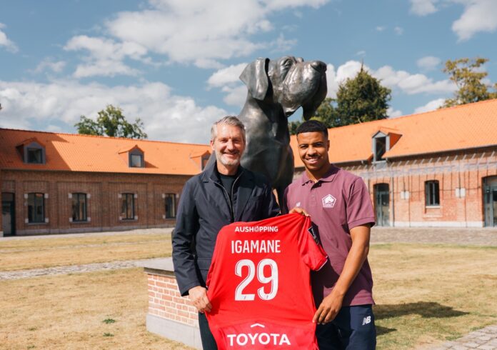 Hamza Igamane poses with a Lille OSC shirt after completing his transfer, standing beside a club official in front of the dog statue at Lille’s training ground.