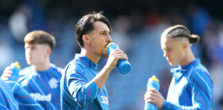 Bojan Miovski takes a drink from a blue water bottle during Rangers warm-up at Ibrox, with team-mates beside him in training kit.