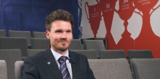 Danny Rohl gives his first interview as Rangers head coach inside Ibrox, smiling confidently in a suit and tie with club trophies shown behind him.