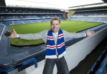 Steven Smith at Ibrox Stadium wearing a Rangers scarf, smiling with arms outstretched overlooking the pitch.
