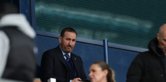 Kevin Thelwell seated in the directors’ area during a Rangers match, appearing deep in thought while holding his phone.
