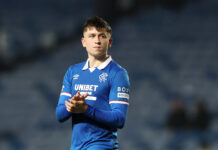 Mikey Moore applauds fans at Ibrox after a Rangers match, wearing the 2025–26 home shirt, highlighting his role as an attacking playmaker.