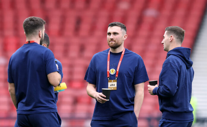 Scotland players in navy training gear stand on the pitch holding cups while talking during a pre match warm up at a stadium with red seats.