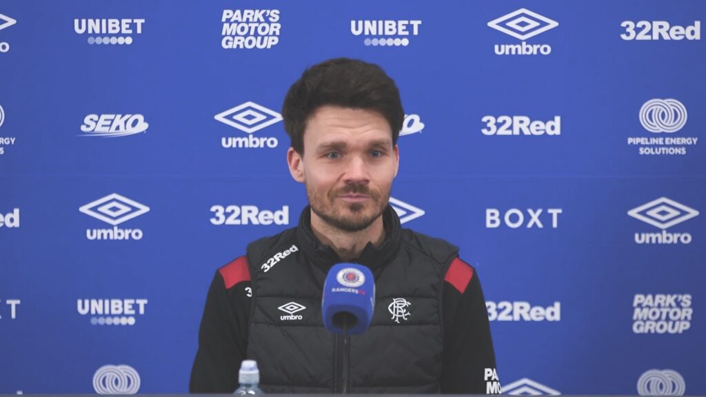 Danny Röhl speaking at a Rangers press conference with Rangers TV microphone in front of blue sponsor backdrop