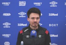 Danny Röhl speaking at a Rangers press conference with Rangers TV microphone in front of blue sponsor backdrop
