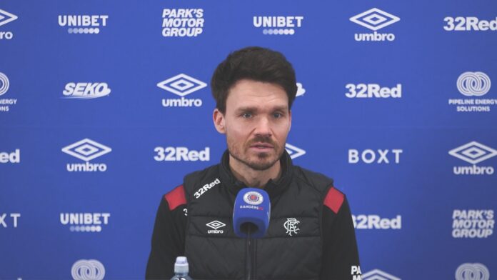 Danny Röhl speaking at a Rangers press conference with Rangers TV microphone in front of blue sponsor backdrop